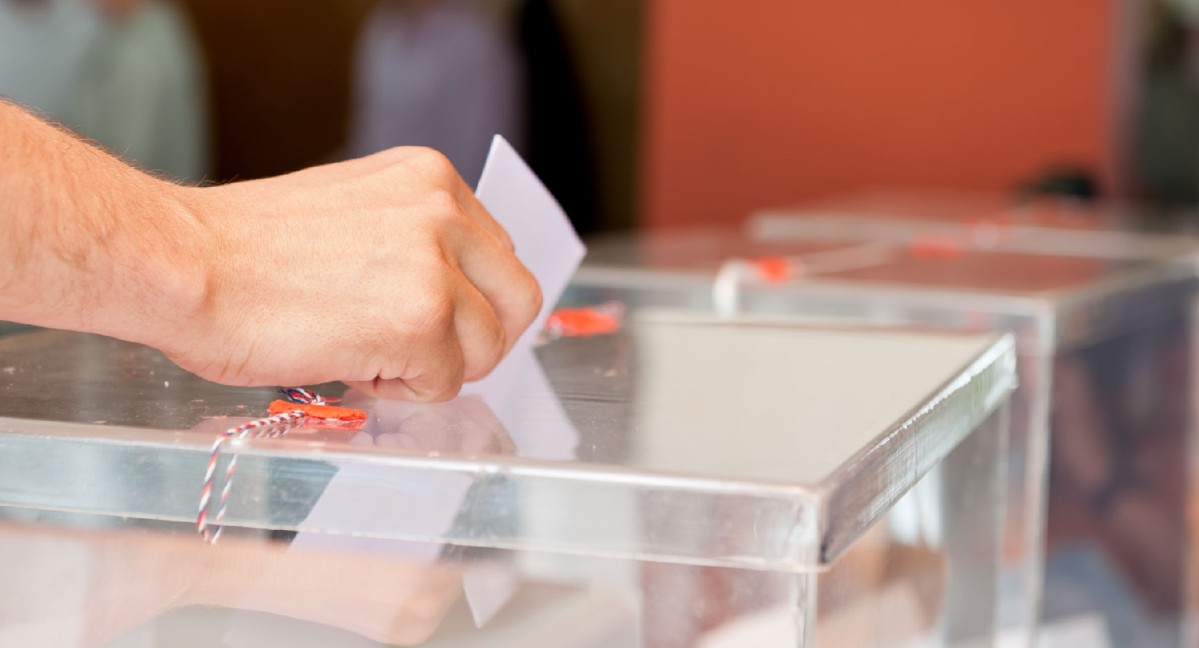 Hand wirft einen Stimmzettel in eine durchsichtige Wahlurne