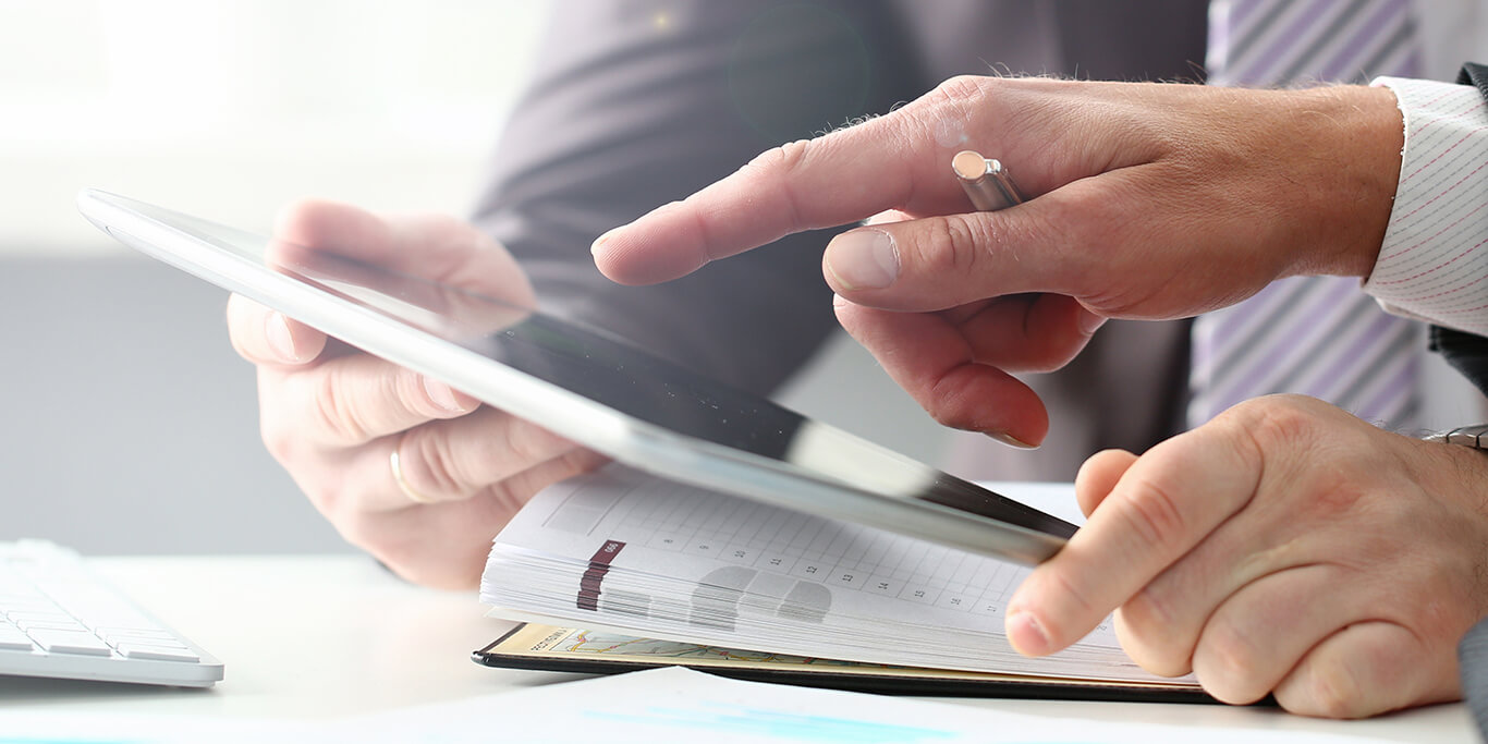 Zwei Personen besprechen Unterlagen, eine Hand zeigt auf den Bildschirm eines Tablets