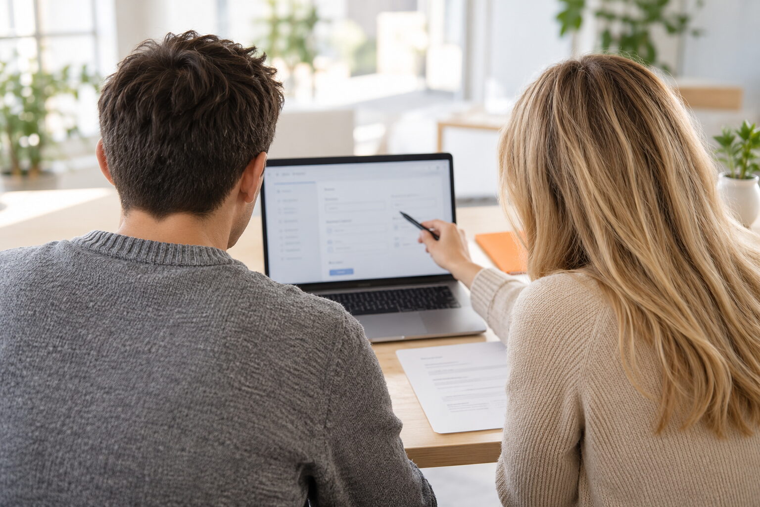 Zwei Personen von hinten an einem Schreibtisch, eine zeigt mit einem Stift auf ein Formular auf dem Laptopbildschirm, daneben liegt ein ausgedrucktes Formular
